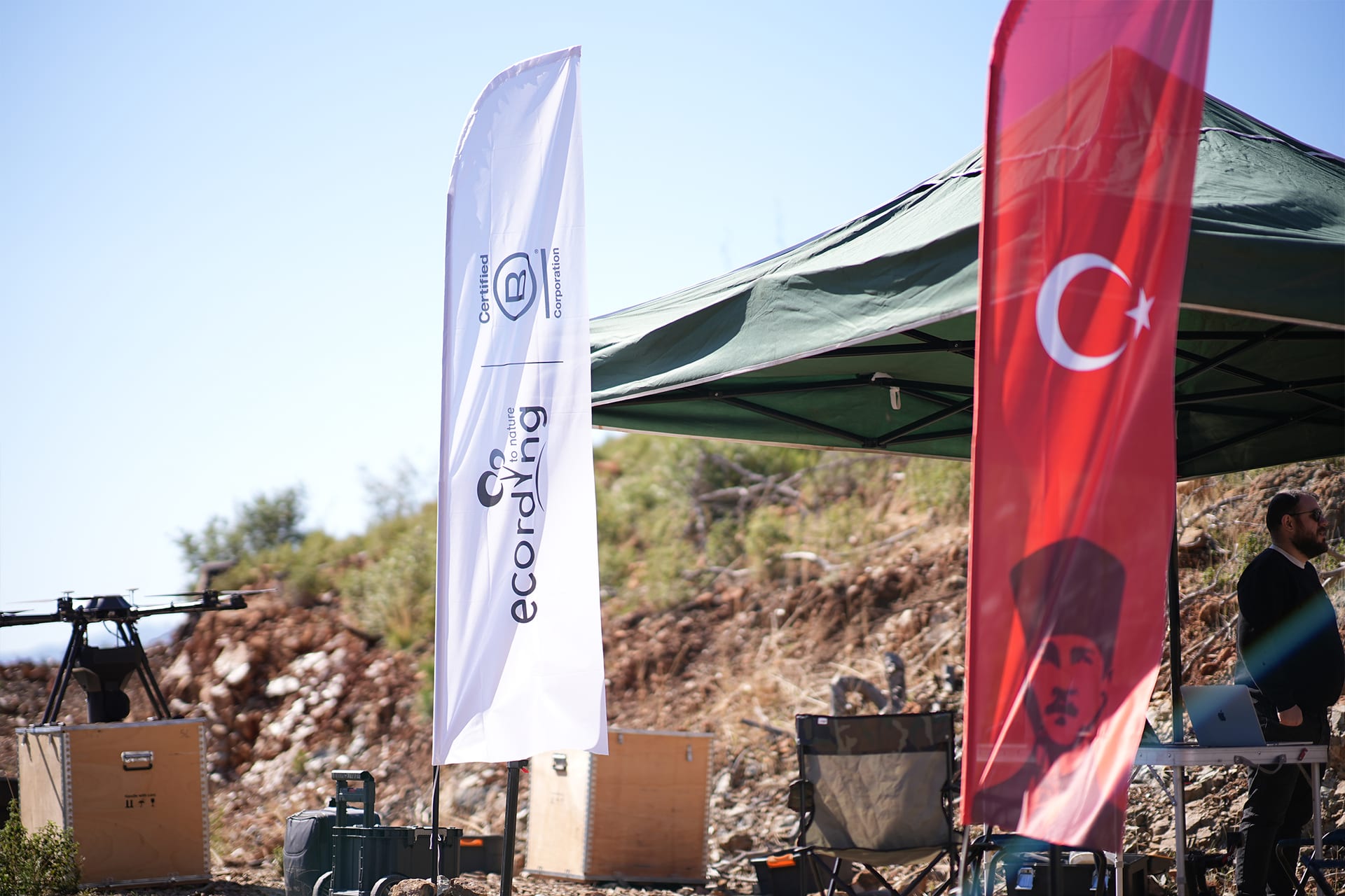 ecording field setup on a hillside, with a branded flag, equipment crates and a drone on the ground